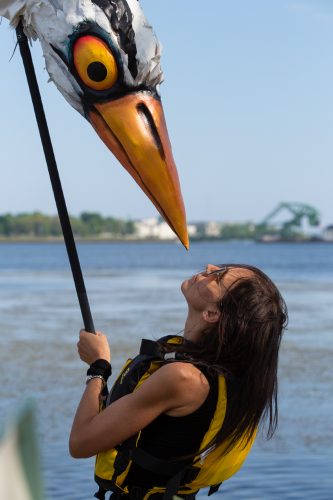 A white woman with straight brown hair, wearing a life jacket, stands in front of the water looking up at the head a large puppet in the form of a great blue heron.