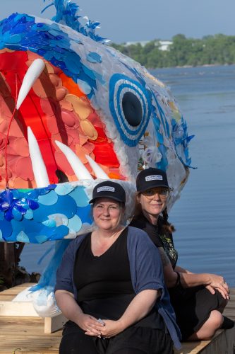 Two white women wearing black and black ball caps sit beside a giant fish puppet created from recycled plastic