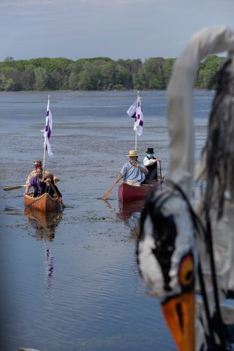 Indigenous passengers in Traditional Clothing paddle a birch bark canoe beside a second canoe made of wood and canvas, containing three people in early 1800s settler attire. Each canoe flies a white and purple two row wampum flag.